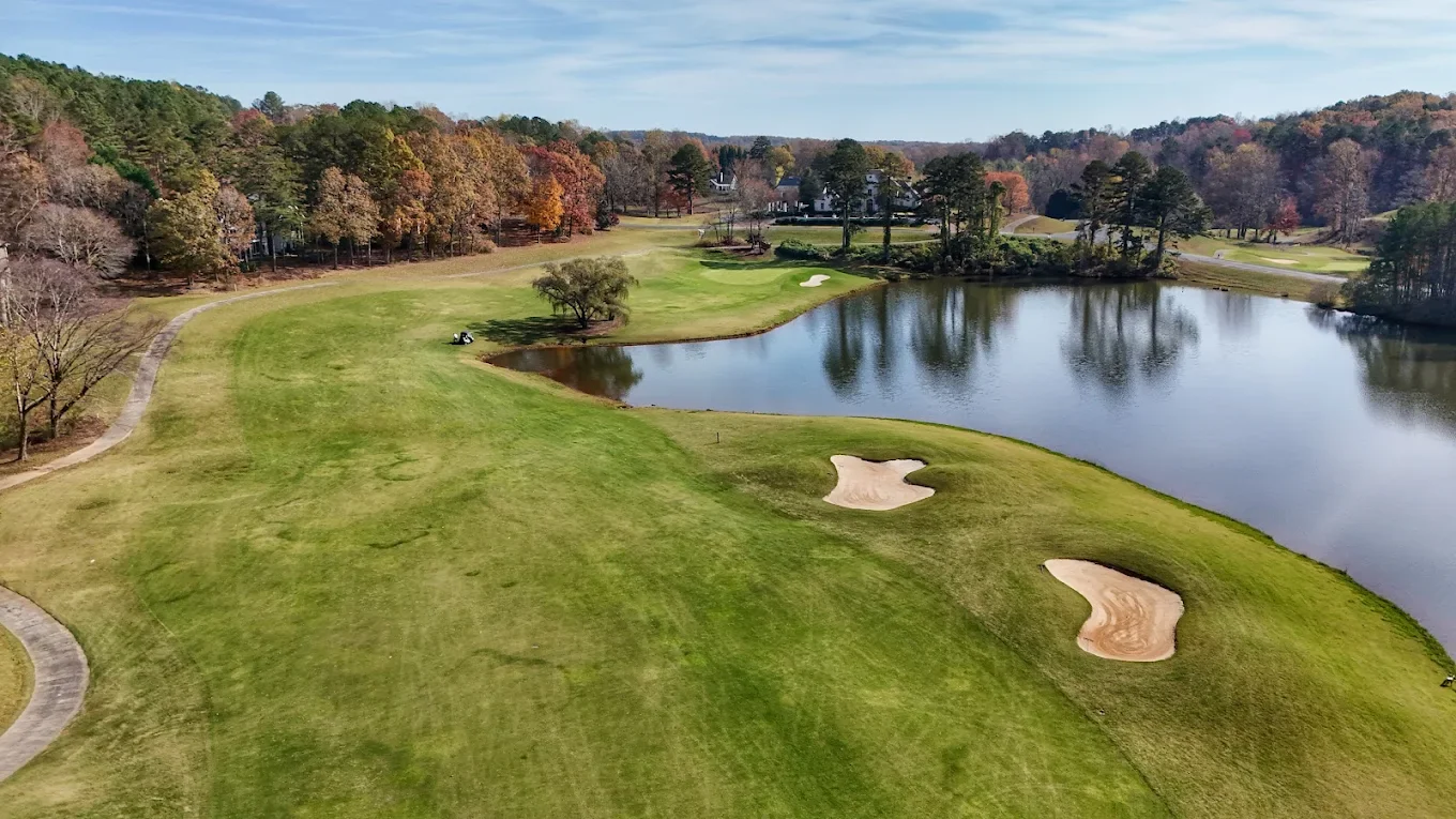 Aerial view of a signature hole at The Orchard, green and sand bunker framed by a scenic pond and autumn hardwoods