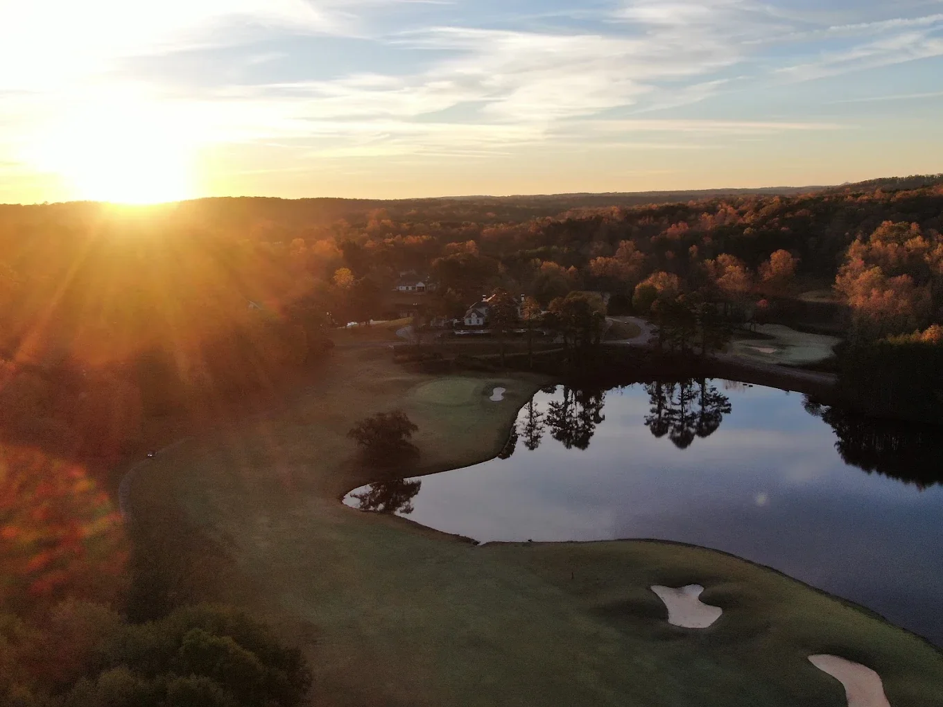 Aerial drone view of The Orchard Golf and Country Club at golden hour, fairways and pond reflecting the sunset